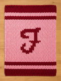A knitted, rectangular throw blanket in pink and burgundy. The top and bottom borders are burgundy, with a burgundy stripe in the middle. A large burgundy letter "J" is centered on the pink field.