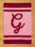 A knitted chunky blanket in shades of pink and burgundy. The top and bottom borders are burgundy, with a lighter pink stripe in between. A burgundy graphic is centered on the pink field.