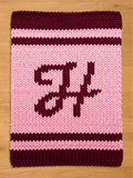 A knitted rectangular blanket in shades of pink and burgundy. The blanket features a large, stylized letter design in burgundy against a pink background. The top and bottom edges are burgundy, with pink and burgundy stripes.