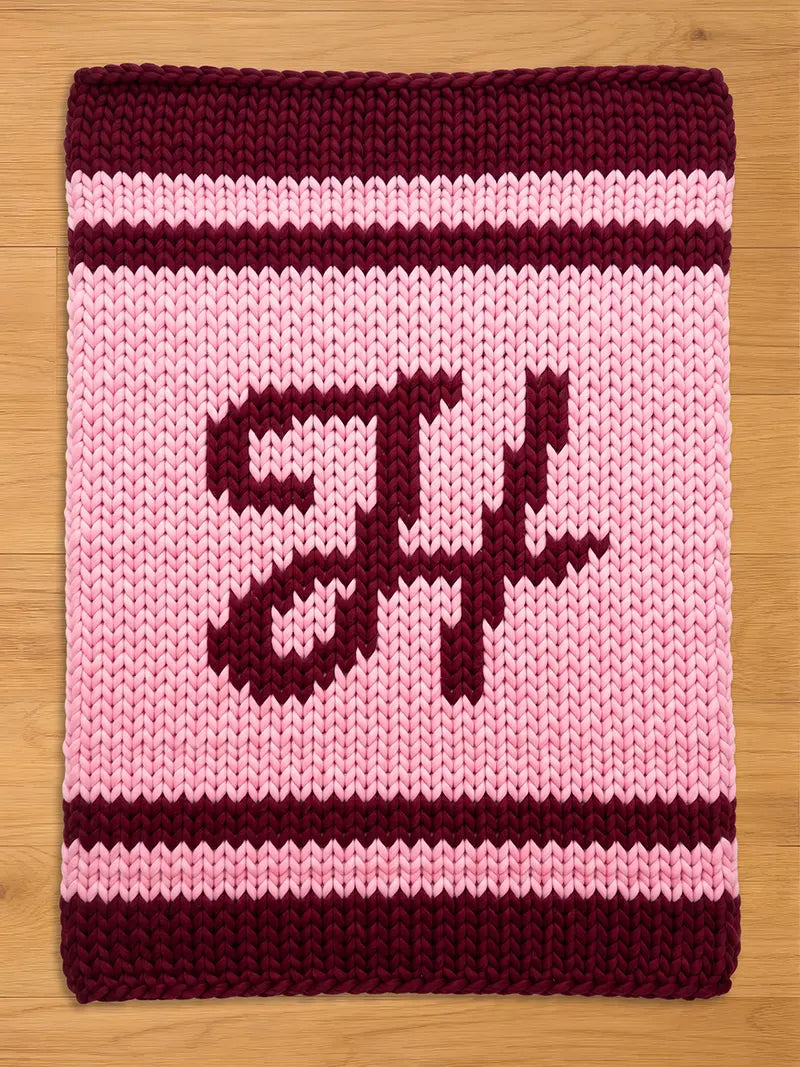A knitted rectangular blanket in shades of pink and burgundy. The blanket features a large, stylized letter design in burgundy against a pink background. The top and bottom edges are burgundy, with pink and burgundy stripes.