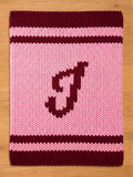 A knitted chunky blanket in shades of pink and burgundy. The blanket features a large letter "I" in burgundy, centered on a pink field. Burgundy stripes border the top and bottom edges, creating a cozy, handmade aesthetic.