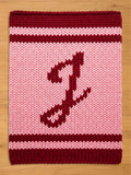 A knitted, rectangular chunky blanket with a pink background and a burgundy letter "J". The top and bottom borders are burgundy, with two burgundy stripes. The item is likely a blanket or pillow.