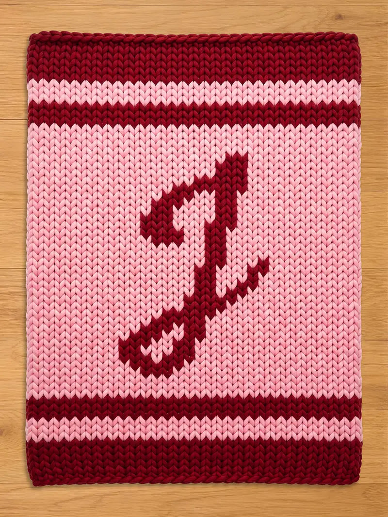 A knitted, rectangular chunky blanket with a pink background and a burgundy letter "J". The top and bottom borders are burgundy, with two burgundy stripes. The item is likely a blanket or pillow.