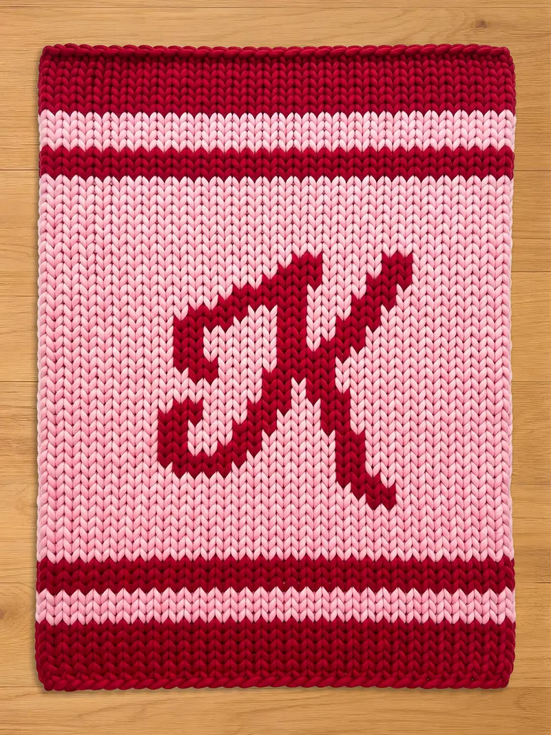 A knitted chunky blanket in shades of red and pink. The top and bottom borders are a deep red, with a lighter red stripe in the middle. The center features a large, stylized letter "K" in a darker red.
