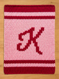 A knitted chunky blanket in shades of red and pink. The top and bottom borders are a deep red, with a lighter red stripe in the middle. The center features a large, stylized letter "K" in a darker red.