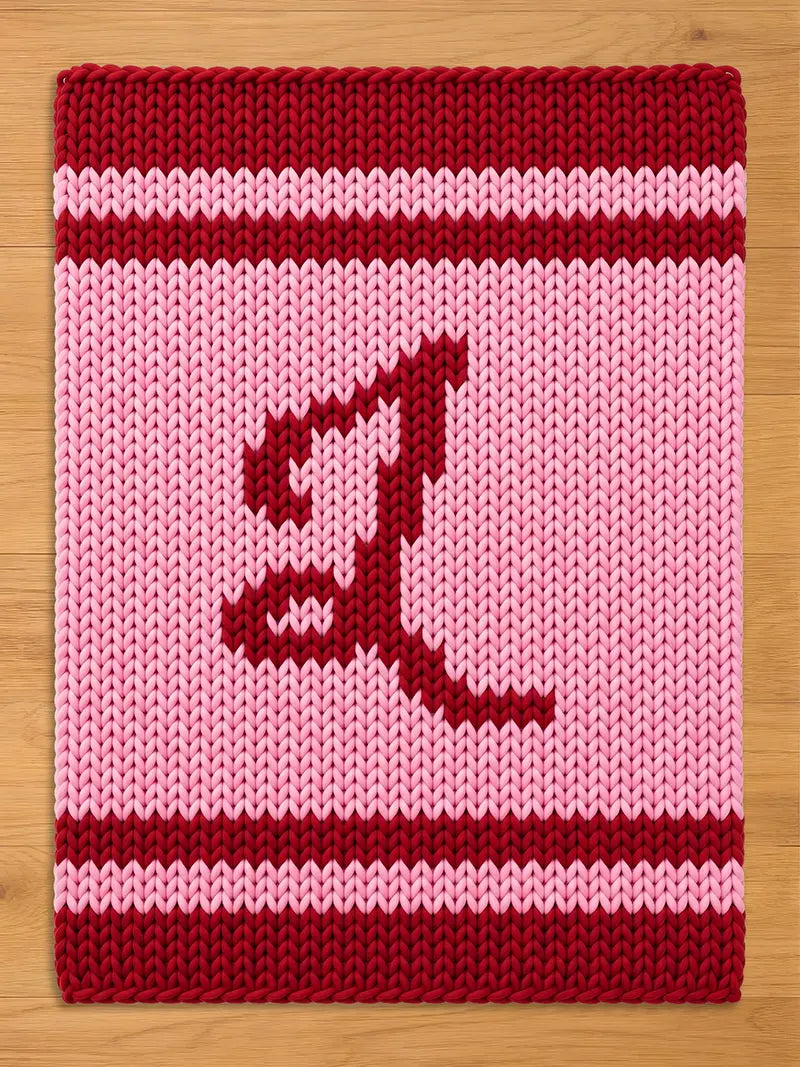 A knitted rectangular item with a pink background and a large, stylized letter "L" in a darker red color. The top and bottom edges are a solid dark red, with a red stripe in the middle.