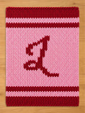 A knitted rectangular item with a pink background and a large, stylized letter "L" in a darker red color. The top and bottom edges are a solid dark red, with a red stripe in the middle.