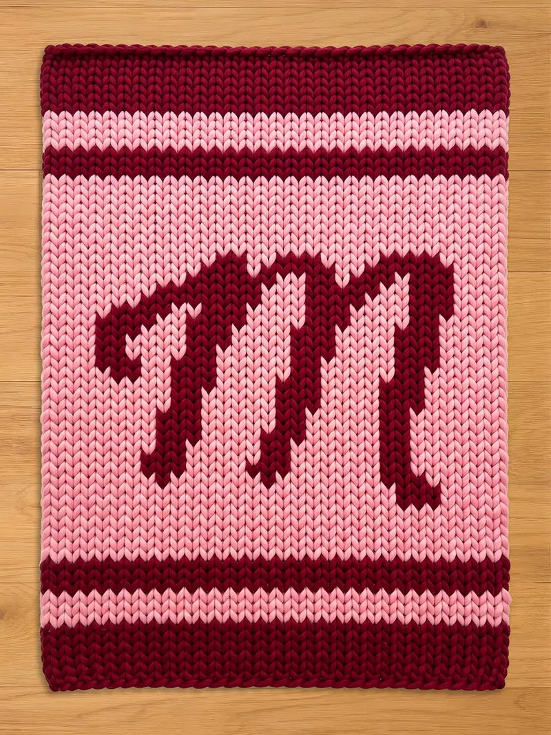 A knitted rectangular chunky blanket in shades of pink and burgundy. The blanket features a large burgundy letter "M" in the center, with burgundy stripes at the top and bottom. The background is a lighter pink.