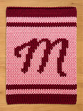 A knitted rectangular chunky blanket in shades of pink and burgundy. The blanket features a large burgundy letter "M" in the center, with burgundy stripes at the top and bottom. The background is a lighter pink.