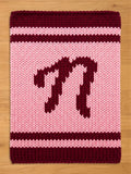 A knitted chunky blanket in shades of pink and burgundy. The central field is light pink, with a burgundy letter "N" in the center. The top and bottom borders are burgundy, and there are burgundy stripes above and below the letter.