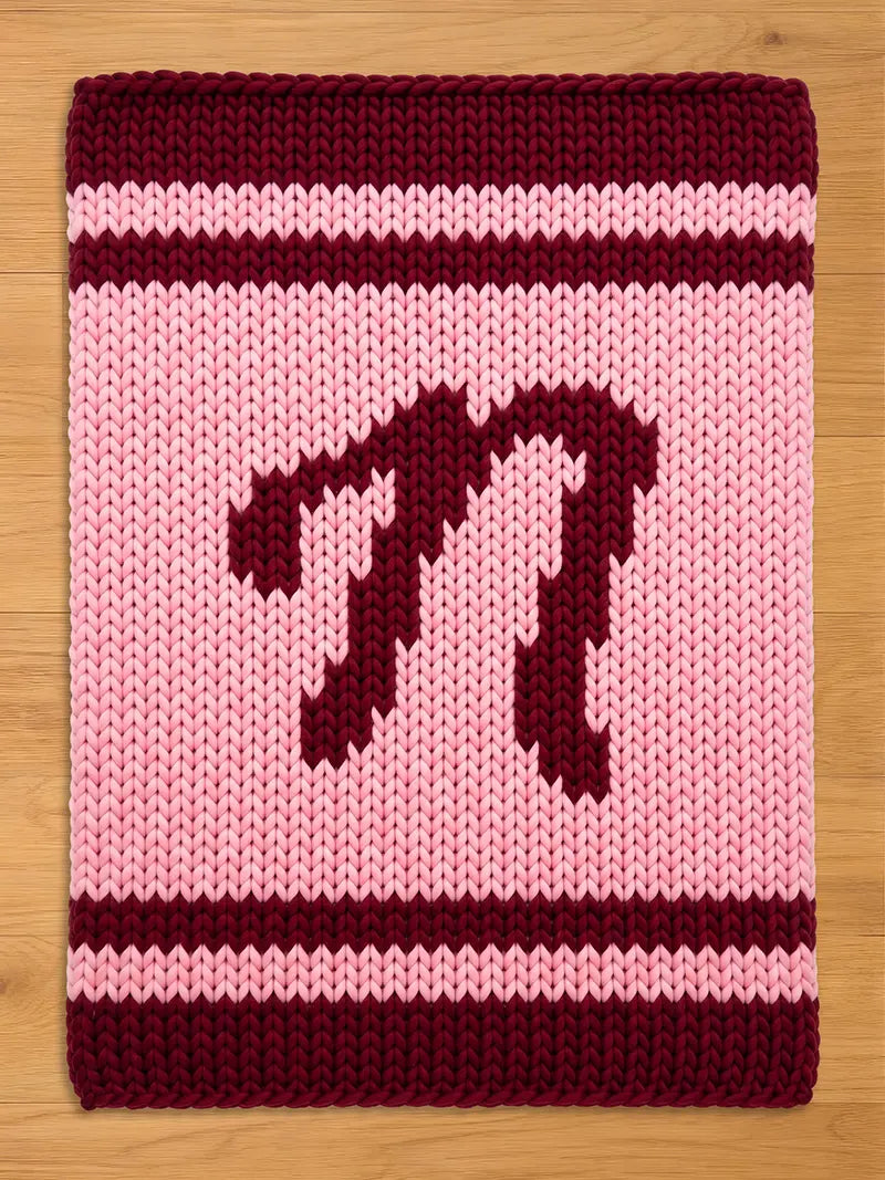 A knitted chunky blanket in shades of pink and burgundy. The central field is light pink, with a burgundy letter "N" in the center. The top and bottom borders are burgundy, and there are burgundy stripes above and below the letter.