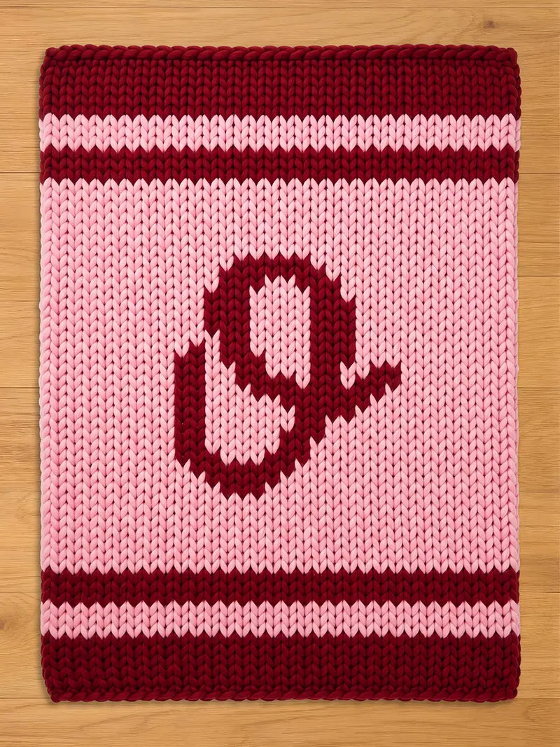 A knitted blanket in shades of pink and burgundy. The blanket features a large letter "O" in burgundy in the center. The top and bottom edges have burgundy stripes, and the background is light pink.