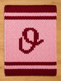 A knitted blanket in shades of pink and burgundy. The blanket features a large letter "O" in burgundy in the center. The top and bottom edges have burgundy stripes, and the background is light pink.