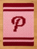 A knitted rectangular item in shades of pink and burgundy. The item features a large burgundy letter "P" in the center. The top and bottom edges have burgundy stripes, and the background is light pink.