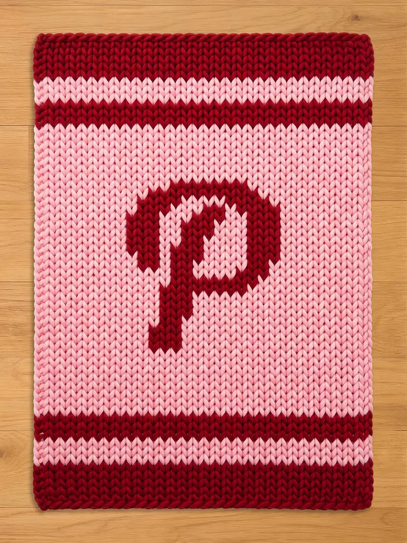 A knitted rectangular item in shades of pink and burgundy. The item features a large burgundy letter "P" in the center. The top and bottom edges have burgundy stripes, and the background is light pink.