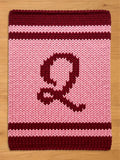 A knitted, rectangular item with a large letter "Q" in burgundy on a pink background. The top and bottom edges feature burgundy stripes, and the overall texture suggests a cozy, handmade feel.