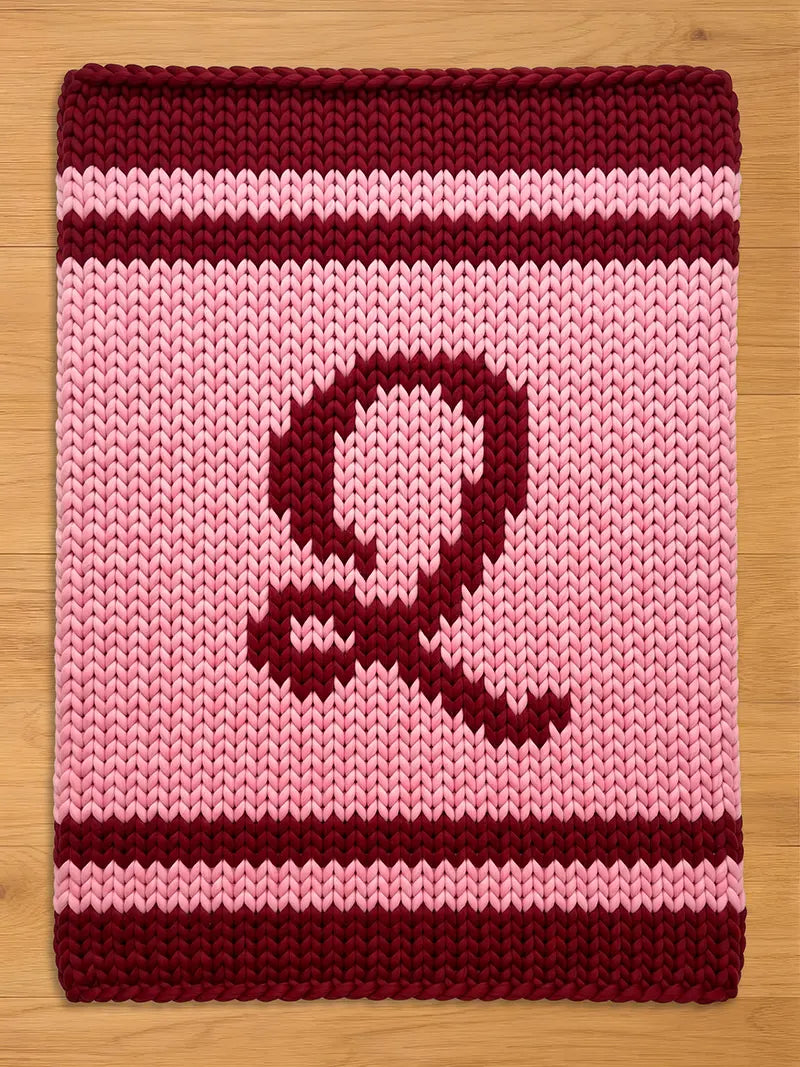 A knitted, rectangular item with a large letter "Q" in burgundy on a pink background. The top and bottom edges feature burgundy stripes, and the overall texture suggests a cozy, handmade feel.