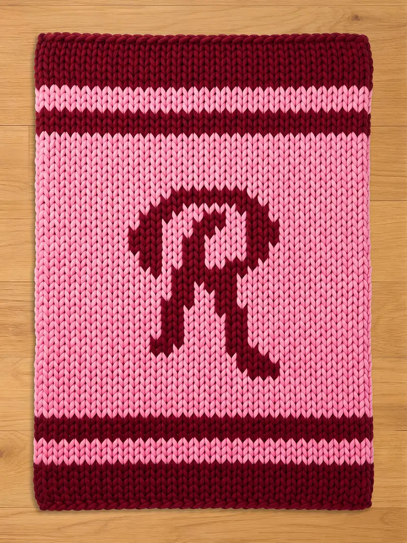 A knitted chunky blanket in shades of pink and burgundy. The blanket features a large letter "R" in burgundy on a pink background. Burgundy stripes border the top and bottom edges. The blanket is made of a thick, chunky knit.