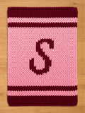  A knitted pink blanket with a burgundy border and stripes. A large burgundy letter "S" is centered on the blanket. The blanket is made of a thick, textured yarn.