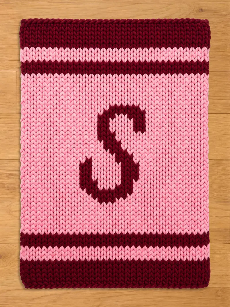  A knitted pink blanket with a burgundy border and stripes. A large burgundy letter "S" is centered on the blanket. The blanket is made of a thick, textured yarn.