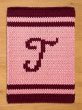 A knitted rectangular chunky blanket with a light pink background and a dark burgundy letter "T". The top and bottom borders are dark burgundy, with a light pink stripe in between. The item is made of thick yarn.
