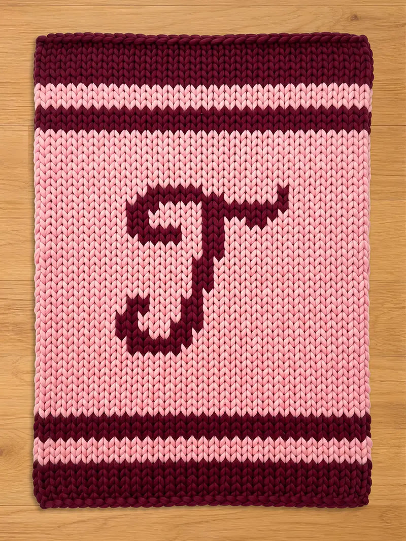 A knitted rectangular chunky blanket with a light pink background and a dark burgundy letter "T". The top and bottom borders are dark burgundy, with a light pink stripe in between. The item is made of thick yarn.