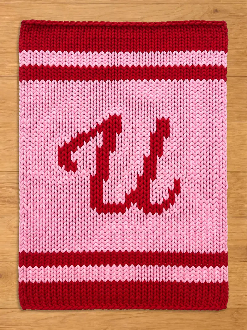 A knitted chunky blanket in shades of red and pink. The border and the letter "W" are in red, while the background and stripes are pink. The "U" is centered on the square.