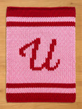 A knitted chunky blanket in shades of red and pink. The border and the letter "W" are in red, while the background and stripes are pink. The "U" is centered on the square.
