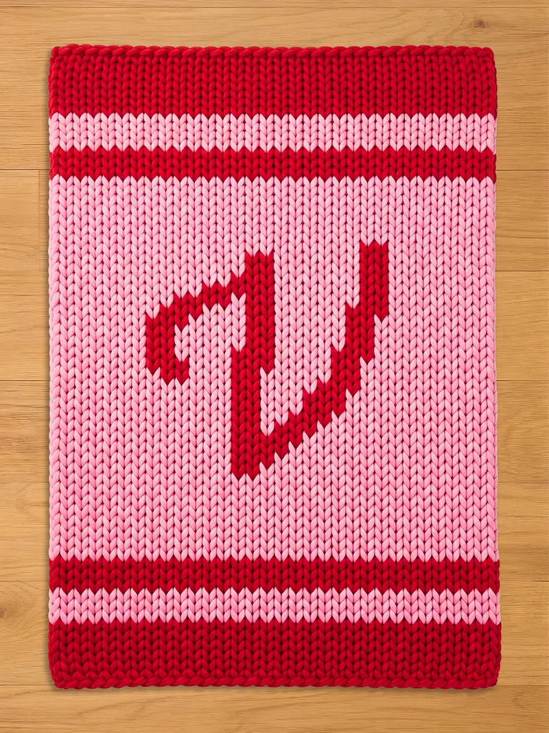 A knitted chunky blanket in shades of red and pink. The top and bottom borders are solid red, with a stripe of white between them and the pink center. A red pixelated letter "V" is in the center.
