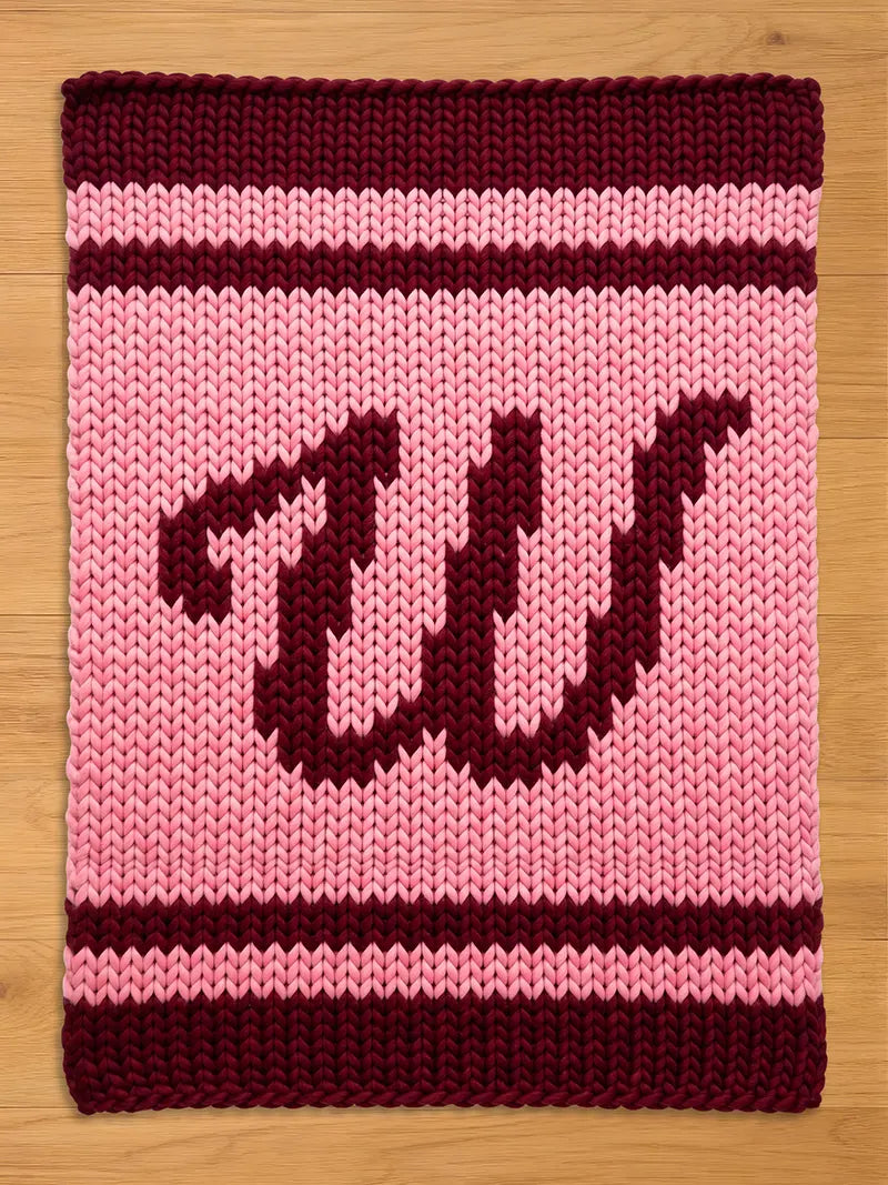 A knitted pink and burgundy chunky blanket with the letters "W" in burgundy. The blanket has a striped border in burgundy and pink. The chunky knit design gives the blanket a cozy, handmade feel.