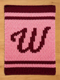 A knitted pink and burgundy chunky blanket with the letters "W" in burgundy. The blanket has a striped border in burgundy and pink. The chunky knit design gives the blanket a cozy, handmade feel.