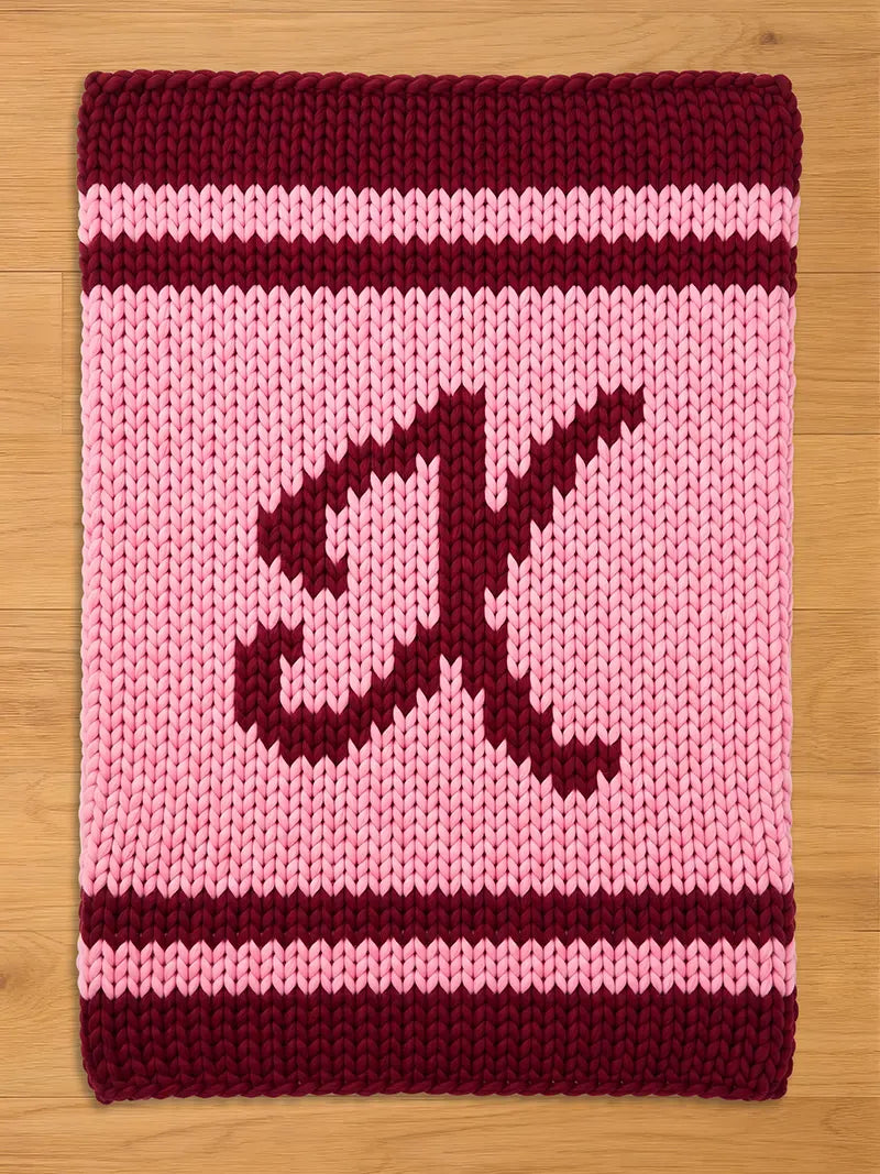 A knitted blanket in shades of pink and burgundy. The blanket features a large letter "X" in burgundy, centered on a pink field. Burgundy stripes border the top and bottom edges. The blanket is made of thick yarn.