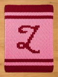 A knitted pink and burgundy blanket with the letter "Z" in burgundy. The blanket has a thick, textured appearance, with the letter centered on the pink field. Burgundy stripes border the top and bottom.
