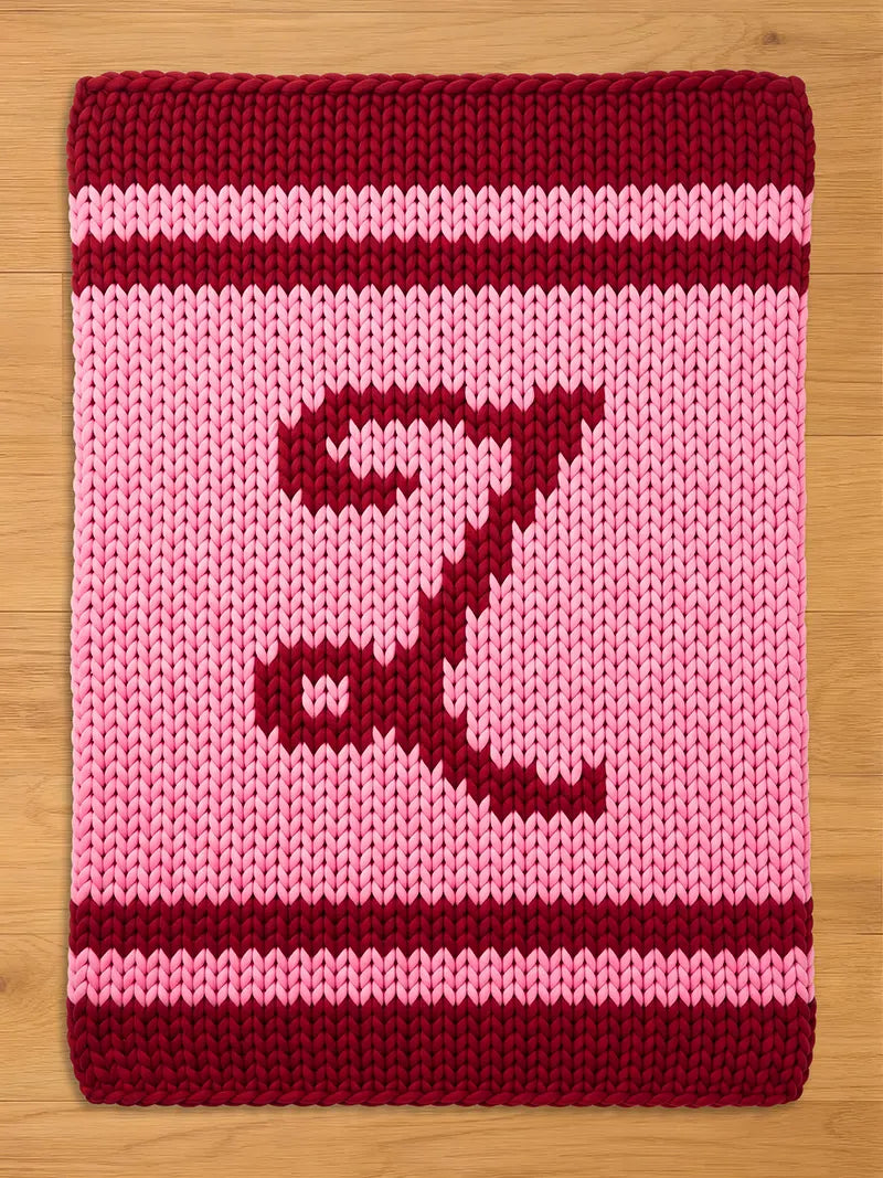 A knitted pink and burgundy blanket with the letter "Z" in burgundy. The blanket has a thick, textured appearance, with the letter centered on the pink field. Burgundy stripes border the top and bottom.