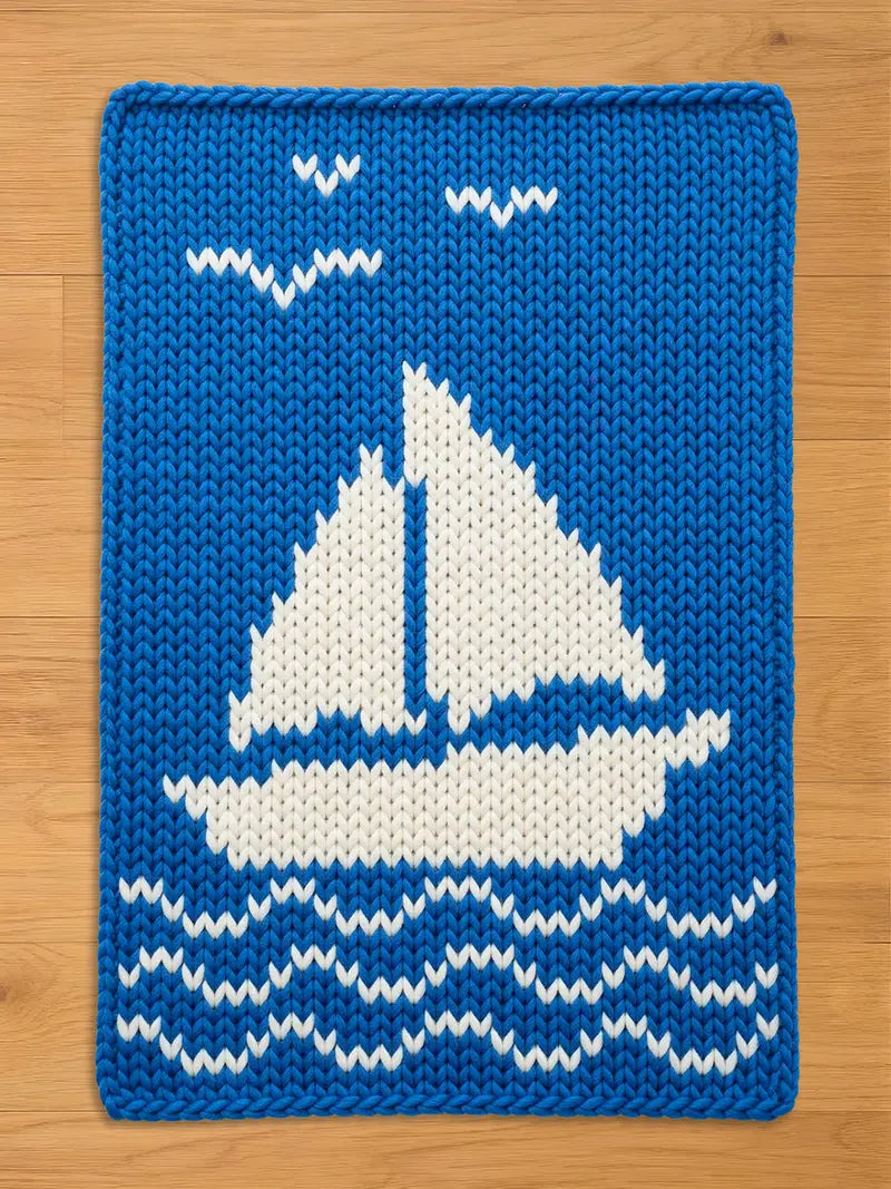 A bright blue chunky knit blanket featuring a white sailboat pattern in the center, with wavy white water lines below and three white birds flying above