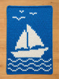 A bright blue chunky knit blanket featuring a white sailboat pattern in the center, with wavy white water lines below and three white birds flying above