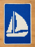 A chunky knit blanket featuring a large white sailing yacht motif on a vibrant blue background, finished with a white border