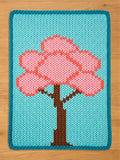 A rectangular chunky knit blanket laid flat on a light wood surface. The blanket features a light blue background with a dark teal border, displaying a large, textured knit pattern of a cherry blossom tree with a brown trunk and vibrant pink blossoms.