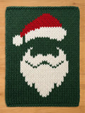 A rectangular knitted blanket featuring a dark green background with a central graphic of a Santa Claus face. The design is created with white yarn for the beard and hat trim, and red yarn for the top of the Santa hat, all in a chunky knit.