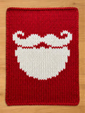 A red chunky knit blanket featuring a large, white knitted design of a Santa Claus beard and curled mustache in the center.