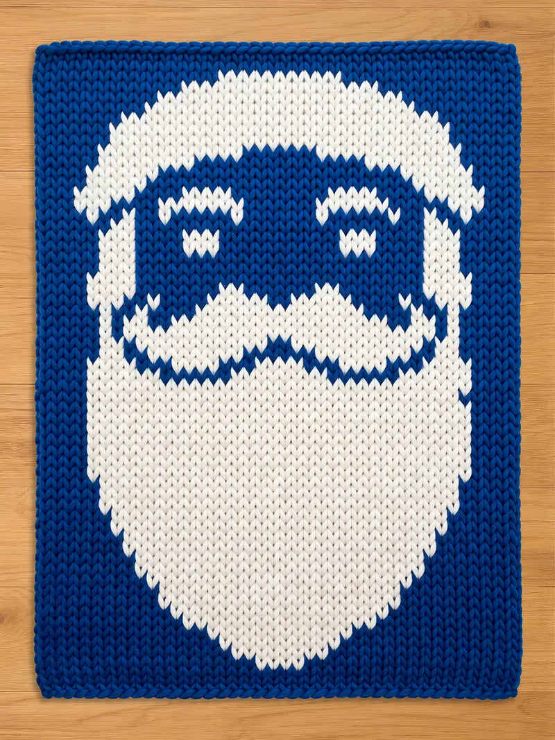 A chunky knitted blanket with blue and white Santa Claus face design. The Santa face features a white beard, mustache, eyebrows, and a blue background. The knitted texture is visible throughout the design.