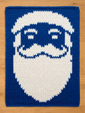 A chunky knitted blanket with blue and white Santa Claus face design. The Santa face features a white beard, mustache, eyebrows, and a blue background. The knitted texture is visible throughout the design.
