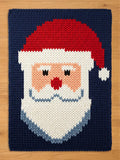 A rectangular chunky knit blanket laid flat on a wooden surface. The blanket features a large, knitted pattern of a Santa Claus head with a red hat, fluffy white beard, and red nose, set against a dark blue background
