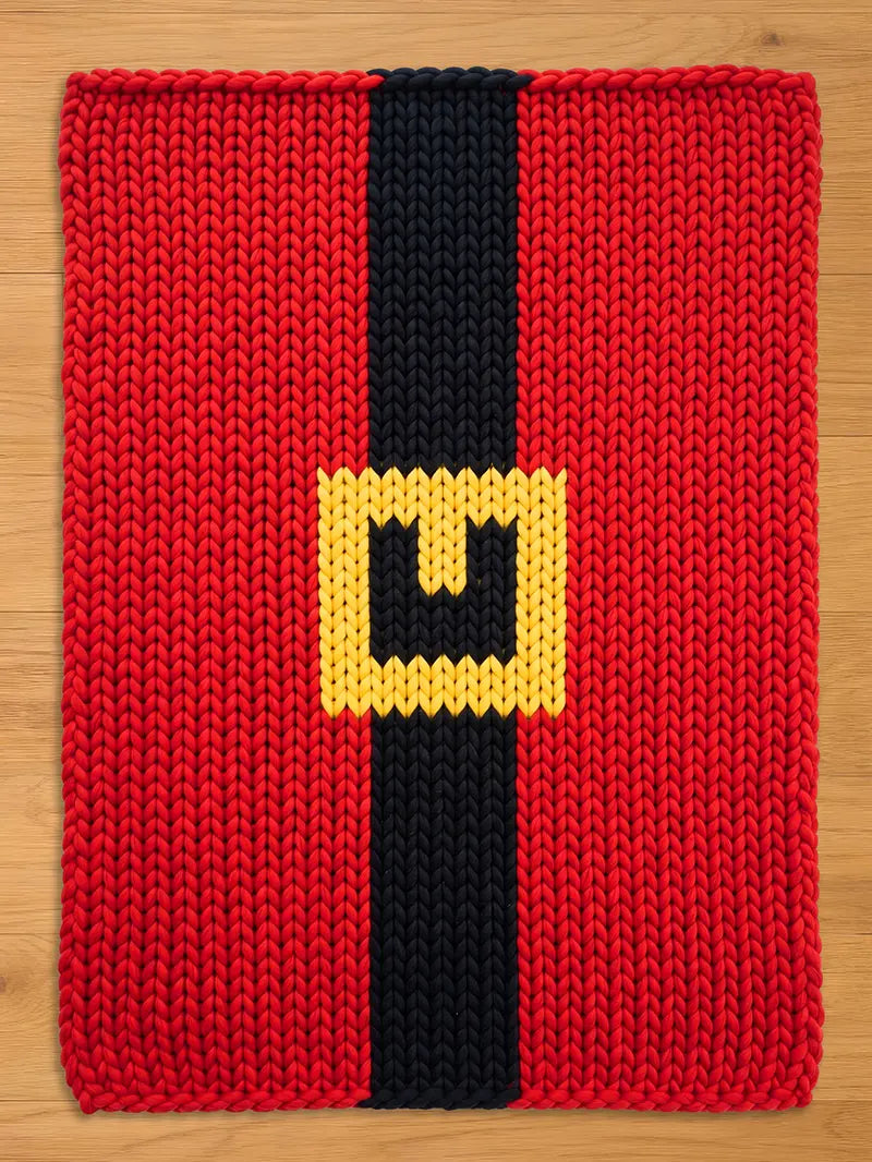 A knitted chunky blanket witth a black vertical stripe and a yellow square buckle. The design resembles Santa Claus's belt. The item is likely a decorative piece or a holiday-themed accessory.