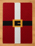 Red chunky knit blanket with a festive Santa suit design. The pattern includes a thick white center stripe and a horizontal black belt with a gold-colored knit buckle, laid flat on a light wood floor.