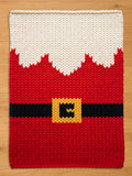 A knitted Christmas-themed chunky blanket featuring a Santa Claus suit design. The top portion is white, mimicking a beard, transitioning to a red body with a black belt and a gold buckle. The design is made of thick yarn.