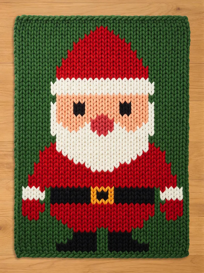 A rectangular knitted chunky blanket featuring a pixelated Santa Claus design. The figure has a red hat with a white trim, a peach face, a white beard, a red coat, a black belt with a yellow buckle, and black boots on a green background.