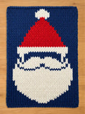A knitted Christmas-themed chunky blanket featuring a Santa Claus face. The design includes a red hat with a white pom-pom, a white beard, and a blue background. The rug is rectangular and made of thick yarn.