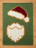 A rectangular knitted blanket featuring a chunky olive green background with a central graphic design of a white Santa Claus beard and a red Santa hat with a white trim, all constructed with a thick, textured yarn knit pattern.