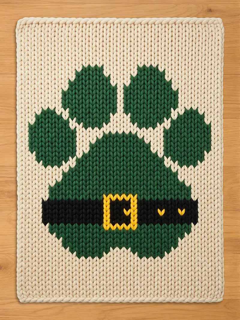 A rectangular chunky knit blanket in cream featuring a large green paw print design. The paw print includes a black horizontal band across the bottom section with a yellow rectangular buckle detail in the center of the band.